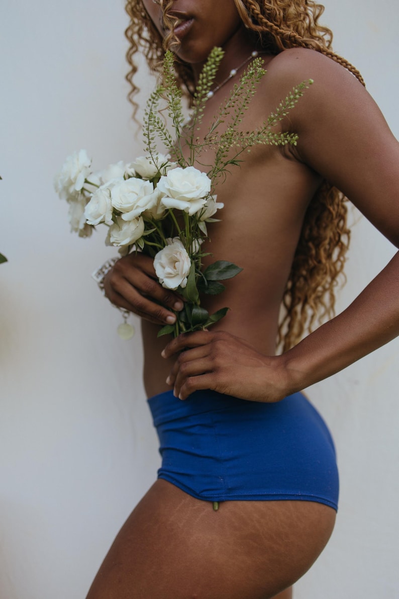 May include: A person holding a bouquet of white roses and greenery, wearing blue shorts. The person's skin is visible, and they are standing in front of a white wall. The image has a natural, soft aesthetic.