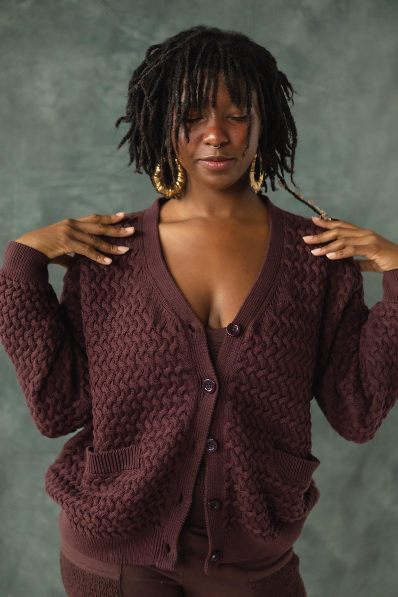 May include: A woman wearing a brown knitted cardigan with a cable knit pattern and a v-neck. The cardigan has two pockets and is buttoned up the front.