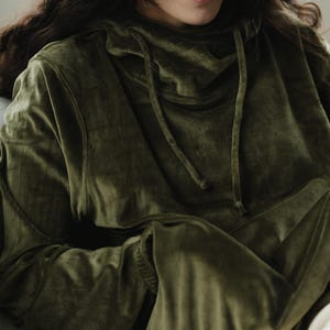 May include: An olive green velvet hoodie with a drawstring neckline. The long-sleeved top has a relaxed fit and a soft, textured appearance. The hoodie is a solid colour and appears comfortable.