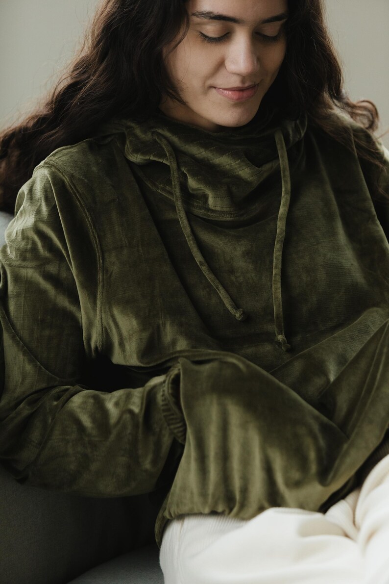 May include: An olive green velvet hoodie with a drawstring neckline. The long-sleeved top has a relaxed fit and a soft, textured appearance. The hoodie is a solid colour and appears comfortable.