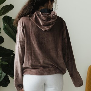 May include: A brown hooded sweatshirt with long sleeves. The sweatshirt is made of a soft, velvety material. The person is wearing light grey sweatpants. The background includes a large green plant and a yellow chair.