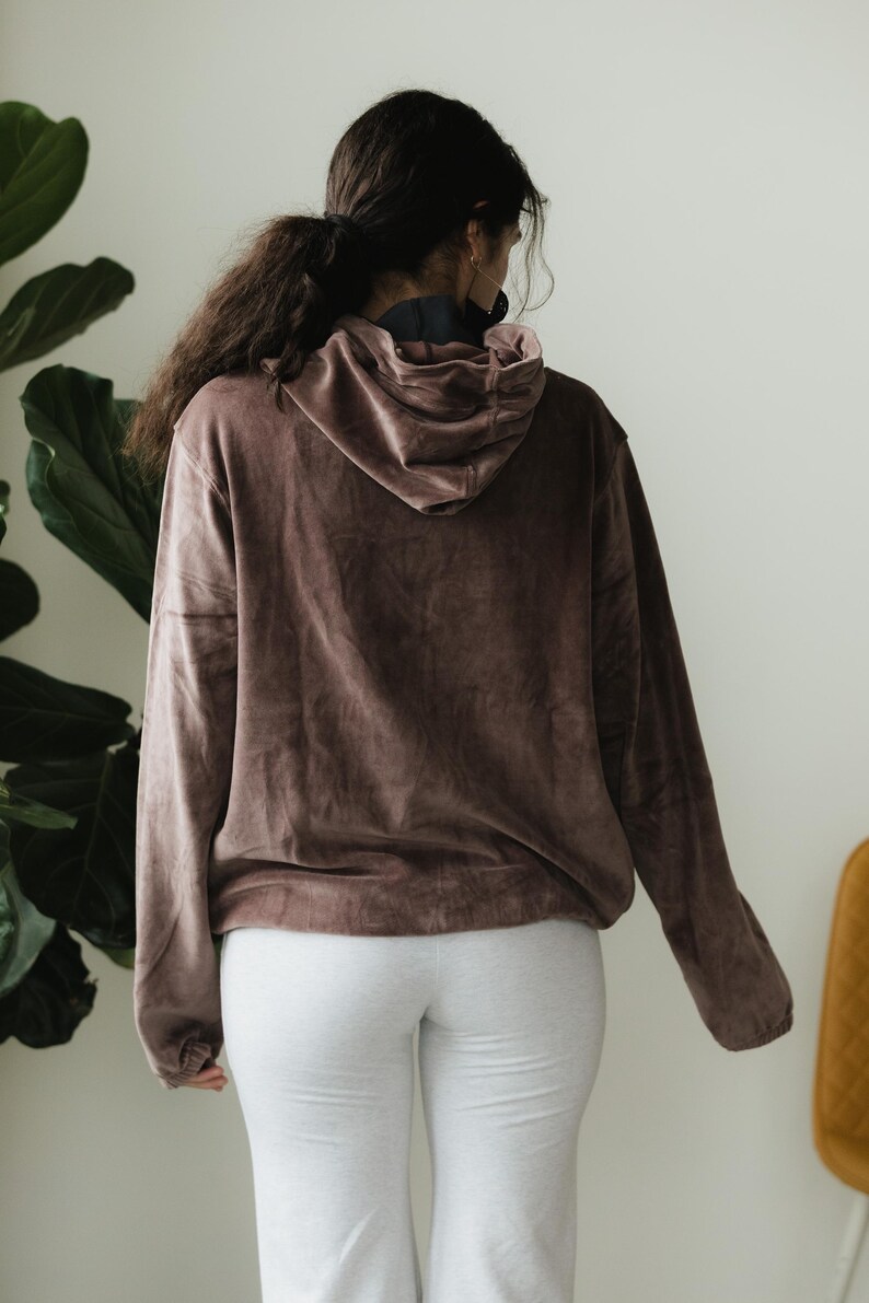 May include: A brown hooded sweatshirt with long sleeves. The sweatshirt is made of a soft, velvety material. The person is wearing light grey sweatpants. The background includes a large green plant and a yellow chair.