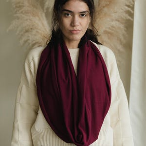 May include: A woman wearing a burgundy infinity scarf over a cream-colored sweater. The scarf is draped around the neck, creating a loop. The background features neutral tones and dried plants.