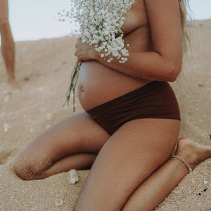 May include: A pregnant person kneeling on a sandy beach, holding a bouquet of white baby's breath flowers. They are wearing a brown bikini bottom.