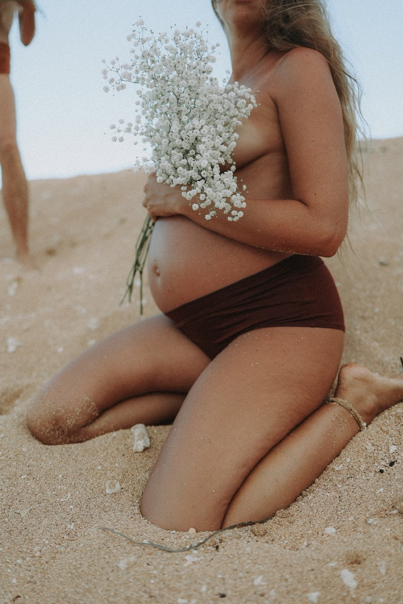 May include: A pregnant person kneeling on a sandy beach, holding a bouquet of white baby's breath flowers. They are wearing a brown bikini bottom.