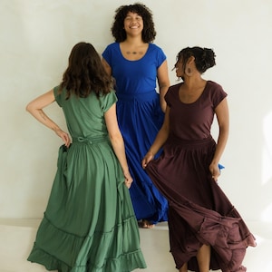 May include: Three women wearing flowing dresses in shades of green, blue, and burgundy. The dresses have short sleeves and tiered skirts. The women are standing on a white surface, with a neutral background.