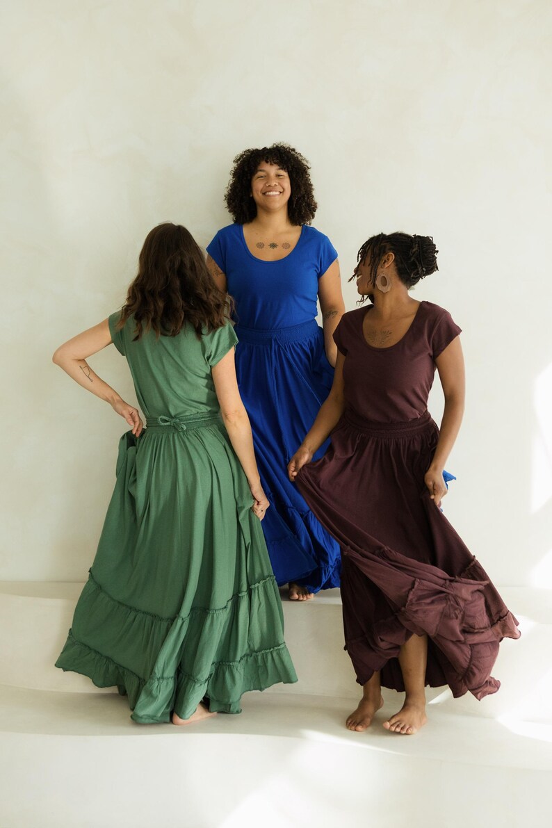 May include: Three women wearing flowing dresses in shades of green, blue, and burgundy. The dresses have short sleeves and tiered skirts. The women are standing on a white surface, with a neutral background.