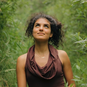 May include: A woman with curly brown hair looks up at the sky. She is wearing a brown cowl neck top and a delicate gold necklace. The background is a lush green forest.