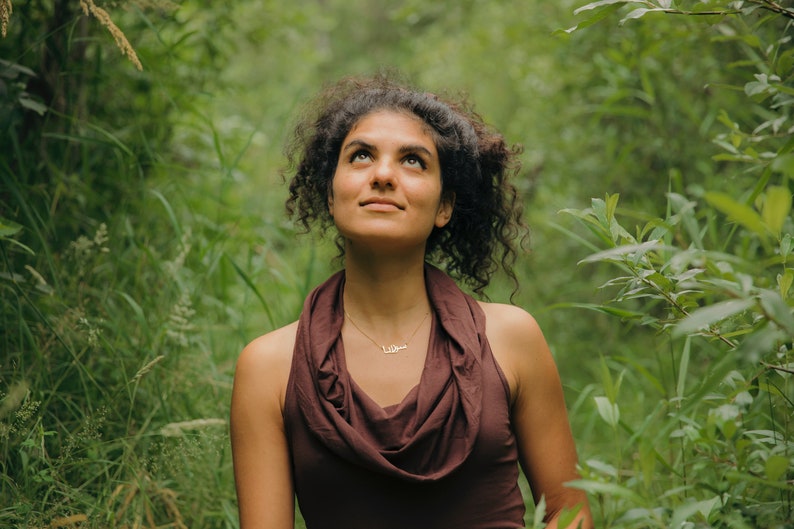 May include: A woman with curly brown hair looks up at the sky. She is wearing a brown cowl neck top and a delicate gold necklace. The background is a lush green forest.