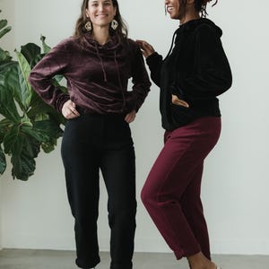 May include: Two people in matching velvet hoodies and trousers. One wears a burgundy hoodie and trousers, the other a black hoodie and trousers. Both wear white trainers. A large green plant is in the background.