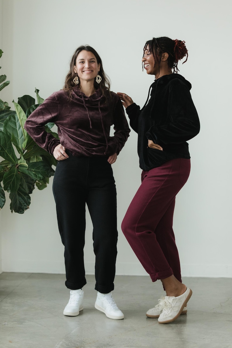 May include: Two people in matching velvet hoodies and trousers. One wears a burgundy hoodie and trousers, the other a black hoodie and trousers. Both wear white trainers. A large green plant is in the background.