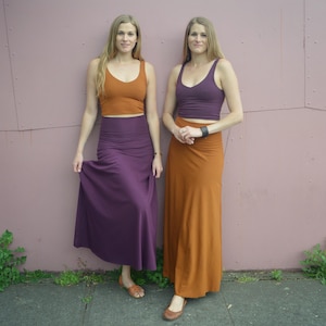 May include: Two individuals wearing matching sets consisting of a solid-colored cropped tank top and a high-waisted maxi skirt. One set is in burnt orange and deep purple, while the other set is in deep purple and burnt orange, both featuring a smooth finish.