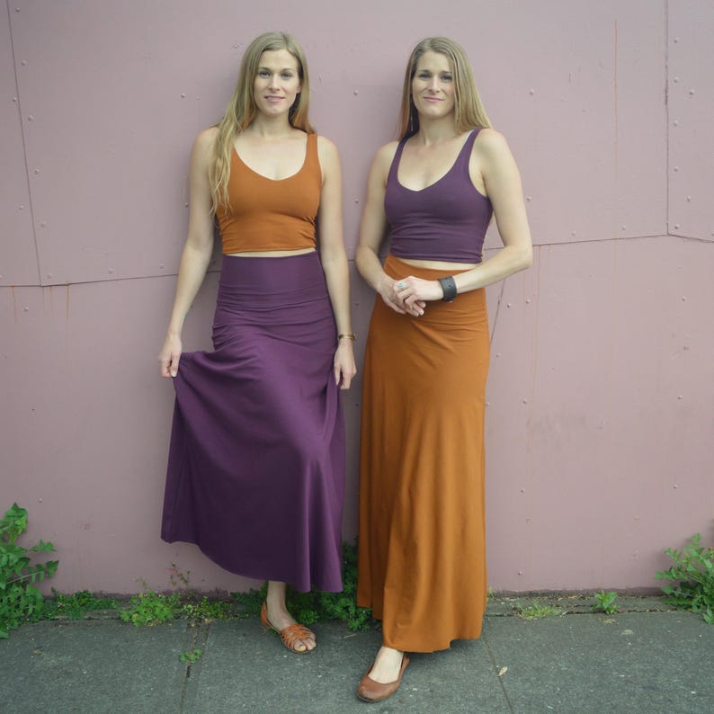 May include: Two individuals wearing matching sets consisting of a solid-colored cropped tank top and a high-waisted maxi skirt. One set is in burnt orange and deep purple, while the other set is in deep purple and burnt orange, both featuring a smooth finish.