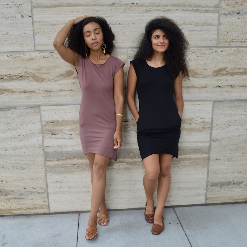 May include: Two short-sleeved dresses, one in a muted mauve color and one in solid black. Both dresses feature a round neckline, a straight-cut silhouette, and visible side pockets. The garments are knee-length with a simple, unadorned design.