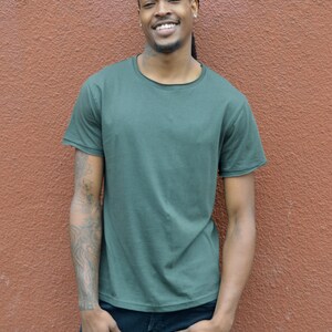 Pu&ograve; includere: Un uomo con i dreadlocks indossa una maglietta verde scuro e jeans blu. Sta sorridendo e guardando la telecamera.