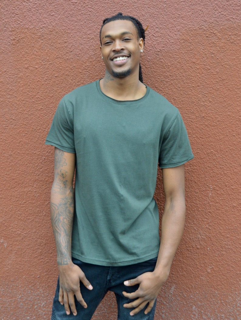 Pu&ograve; includere: Un uomo con i dreadlocks indossa una maglietta verde scuro e jeans blu. Sta sorridendo e guardando la telecamera.