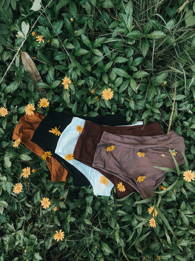 May include: Five pairs of underwear in different colors, including brown, black, white, and purple, are arranged on a bed of green leaves and yellow flowers.