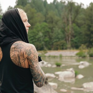 May include: A person with tattoos on their arms and back wearing a black tank top with a hood pulled up over their head. The person is standing in front of a body of water with rocks in the background.