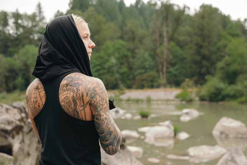 May include: A person with tattoos on their arms and back wearing a black tank top with a hood pulled up over their head. The person is standing in front of a body of water with rocks in the background.