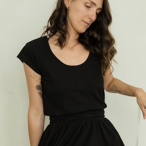 May include: A black scoop-neck t-shirt and a matching black skirt. The skirt has a gathered waistband. The person is wearing gold earrings and a necklace. A tattoo is visible on the left arm.