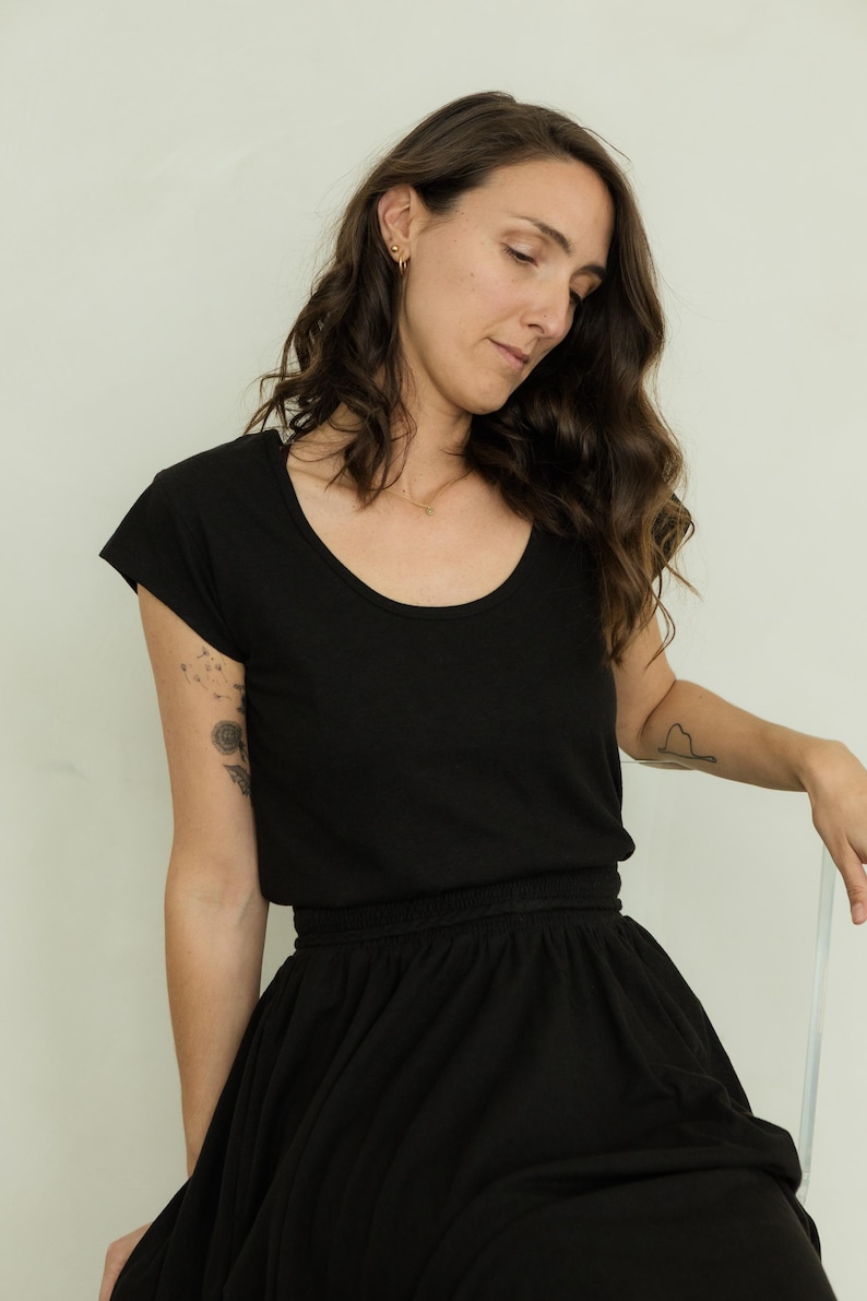 May include: A black scoop-neck t-shirt and a matching black skirt. The skirt has a gathered waistband. The person is wearing gold earrings and a necklace. A tattoo is visible on the left arm.