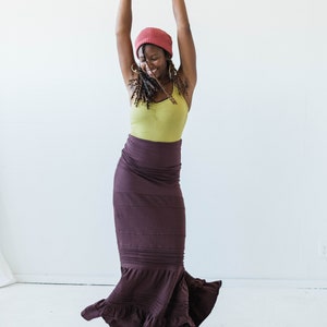 May include: A woman wearing a red beanie, a green tank top, and a long brown skirt with ruffles. She is smiling and has her arms raised in the air.