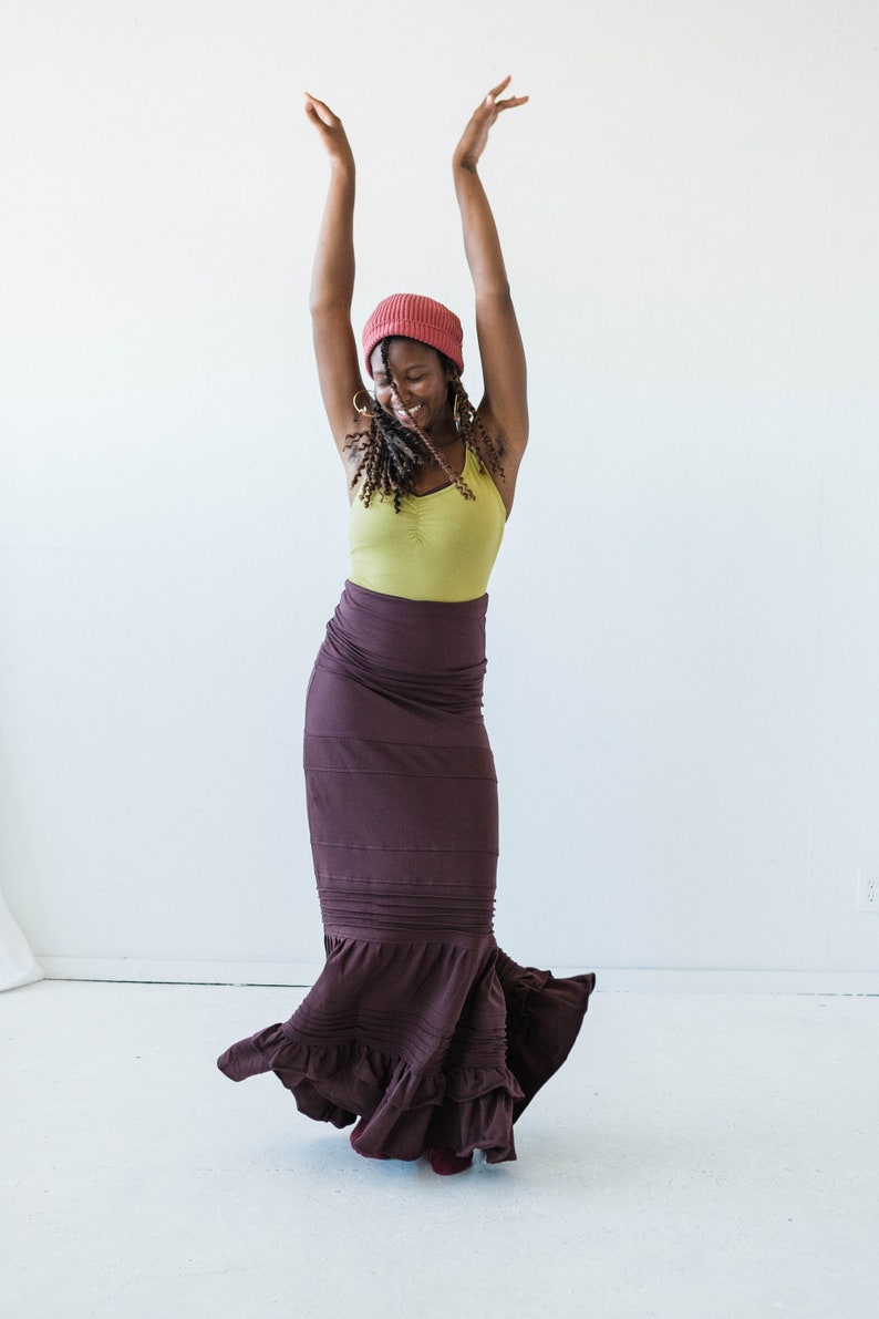 May include: A woman wearing a red beanie, a green tank top, and a long brown skirt with ruffles. She is smiling and has her arms raised in the air.