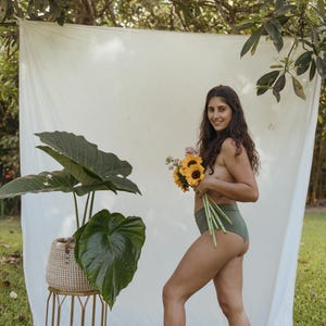 May include: A woman in olive green underwear holds a bouquet of sunflowers and other flowers. A large potted plant with broad green leaves sits on a gold metal stand. A white sheet hangs in the background.