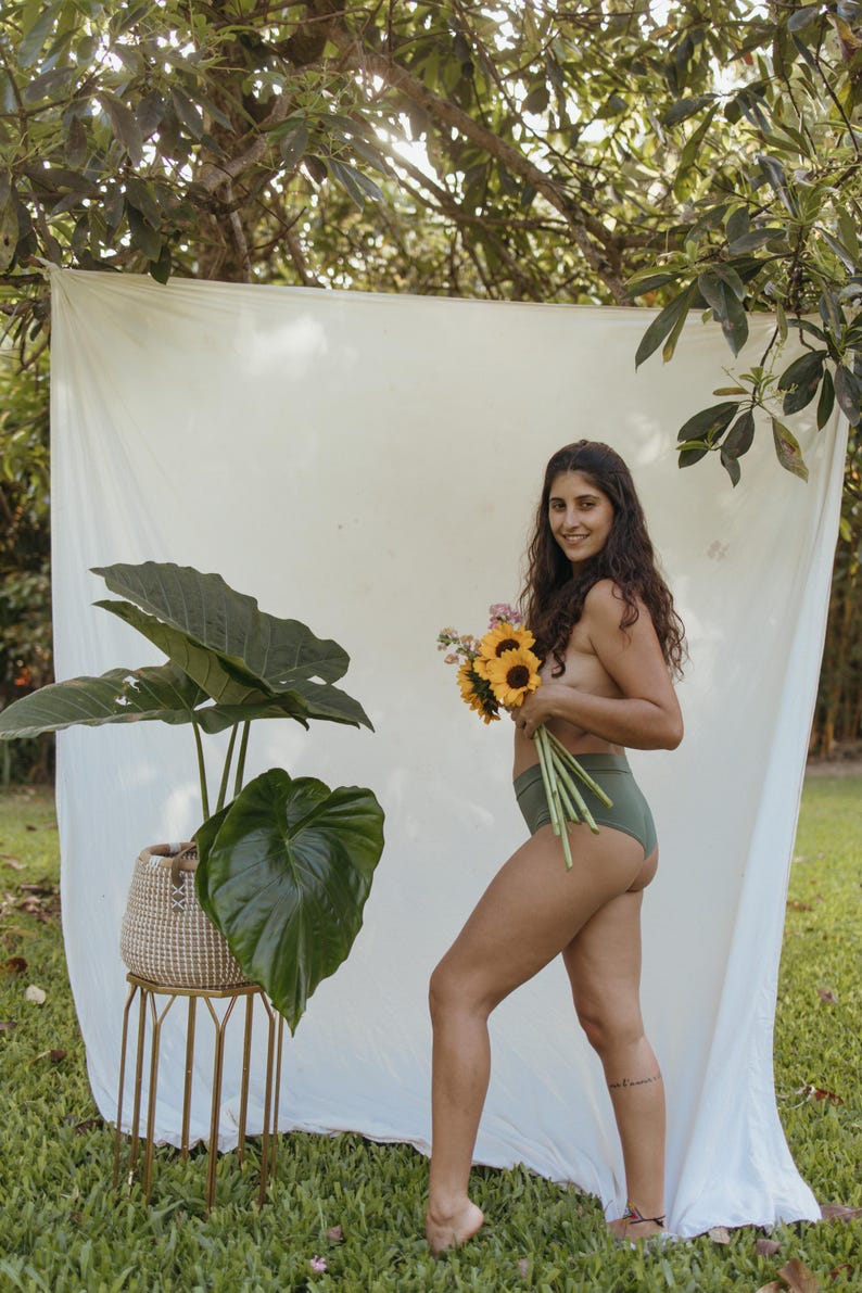 May include: A woman in olive green underwear holds a bouquet of sunflowers and other flowers. A large potted plant with broad green leaves sits on a gold metal stand. A white sheet hangs in the background.