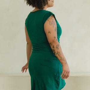 May include: A solid dark green short-sleeved dress with a round neckline and a fitted silhouette that extends to the mid-thigh. The garment features a simple, clean design with a smooth texture and a straight hemline, captured from a side profile view.