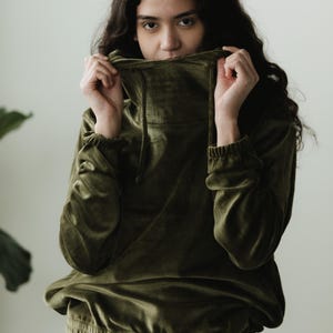 May include: An olive green velvet hoodie with a high neckline and drawstring. The long-sleeved top has elasticated cuffs and a banded waist. The model is holding the neckline up to their face. The background is a plain white wall.