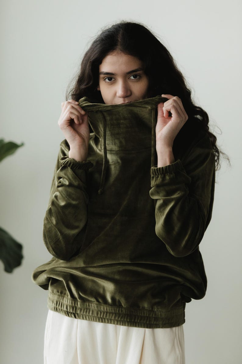 May include: An olive green velvet hoodie with a high neckline and drawstring. The long-sleeved top has elasticated cuffs and a banded waist. The model is holding the neckline up to their face. The background is a plain white wall.