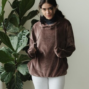 May include: A brown hooded sweatshirt with a drawstring, worn over a black turtleneck, paired with light grey leggings. A large green plant is in the background.