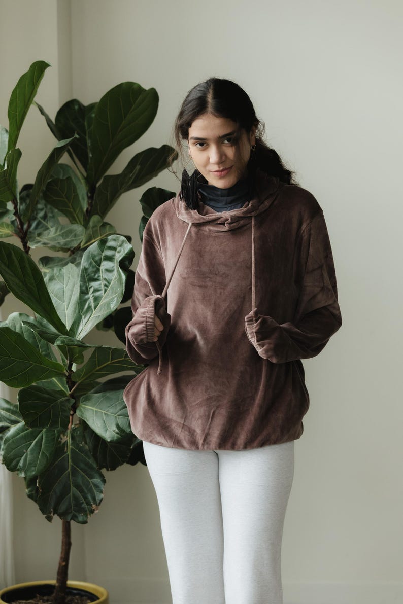 May include: A brown hooded sweatshirt with a drawstring, worn over a black turtleneck, paired with light grey leggings. A large green plant is in the background.