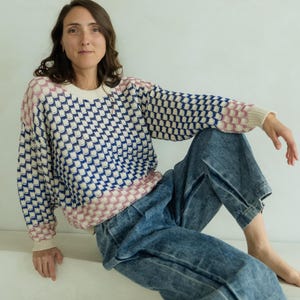 May include: A woman wearing a patterned sweater and denim jeans. The sweater has a white base with a blue and pink geometric design. The jeans are a light wash denim.