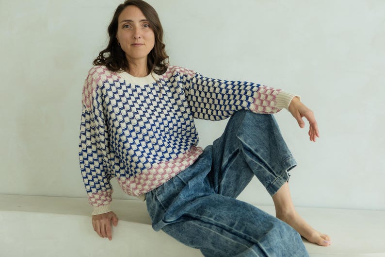 May include: A woman wearing a patterned sweater and denim jeans. The sweater has a white base with a blue and pink geometric design. The jeans are a light wash denim.