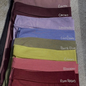 May include: A stack of ten fabric headbands in various colors, including black, earth, cacao, lavender, cornflower, burnt olive, citron, blossom, and rum raisin. Each headband has the color name written on it.