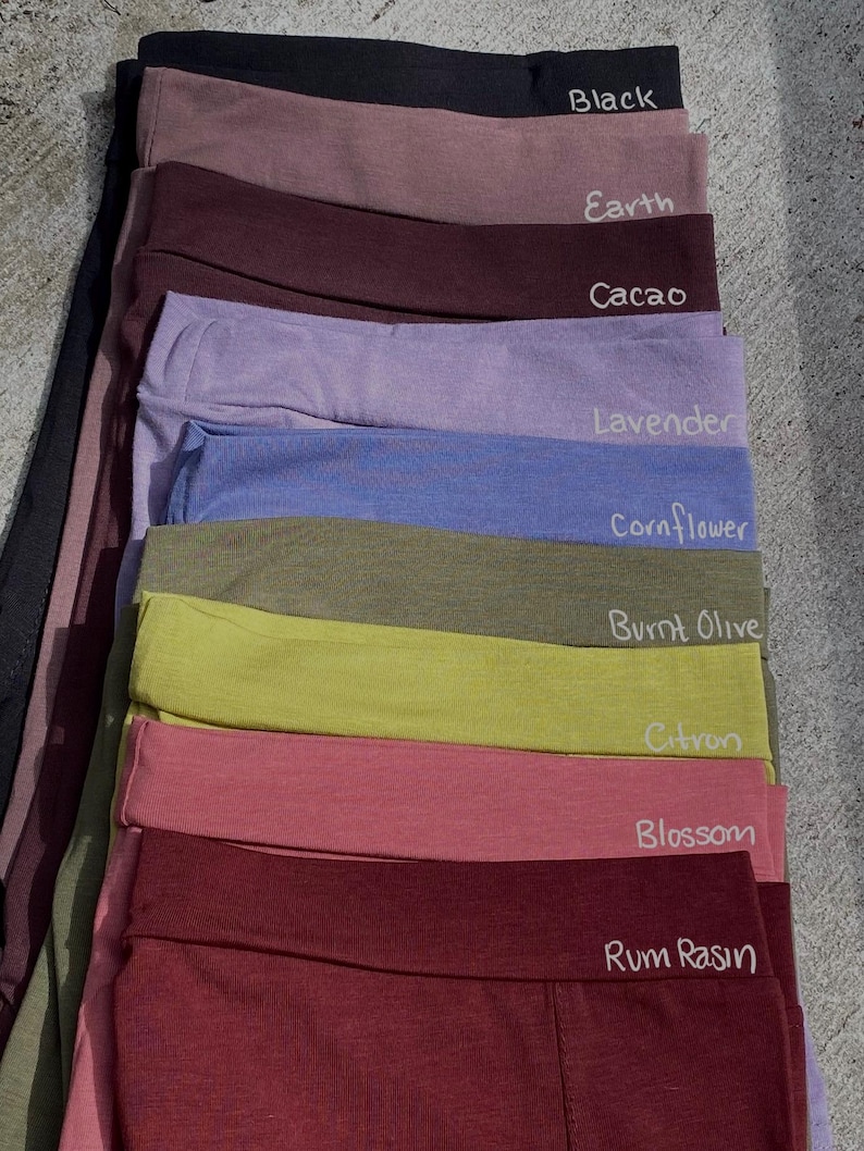 May include: A stack of ten fabric headbands in various colors, including black, earth, cacao, lavender, cornflower, burnt olive, citron, blossom, and rum raisin. Each headband has the color name written on it.