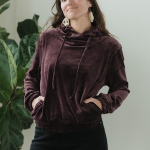 May include: A burgundy velvet hoodie with a drawstring hood and front pockets. The long-sleeved top has a cinched waist and is paired with black trousers. The model is wearing white earrings.