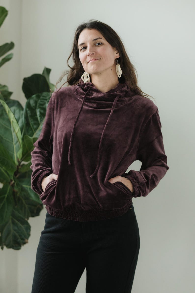 May include: A burgundy velvet hoodie with a drawstring hood and front pockets. The long-sleeved top has a cinched waist and is paired with black trousers. The model is wearing white earrings.
