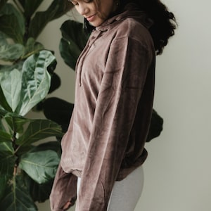 May include: A brown hooded sweatshirt with a drawstring, made of a soft, velvety material. The sweatshirt is paired with a light grey skirt. The model is standing in front of a large green plant.