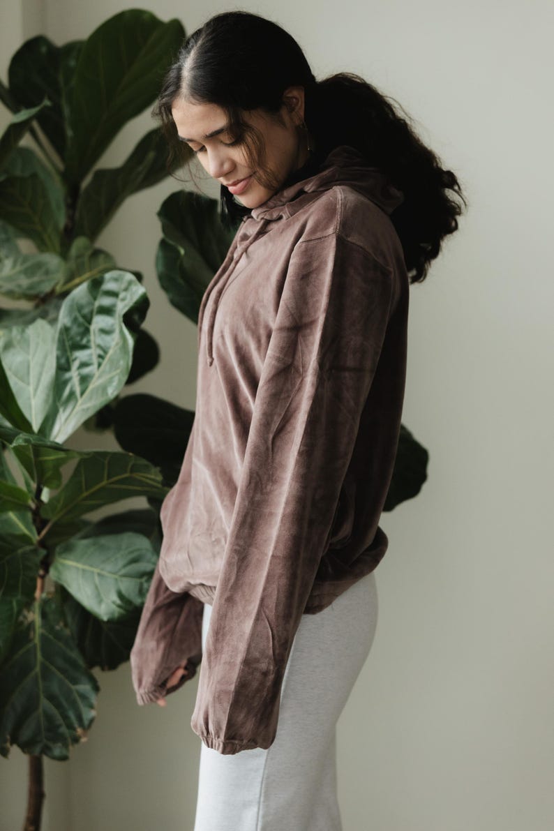 May include: A brown hooded sweatshirt with a drawstring, made of a soft, velvety material. The sweatshirt is paired with a light grey skirt. The model is standing in front of a large green plant.