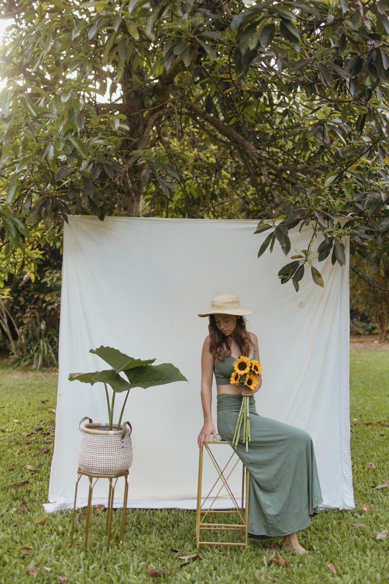 May include: A sage green two-piece set consisting of a cropped tank top and a long, high-waisted skirt. The outfit is paired with a straw hat and a bouquet of yellow sunflowers. A woven basket with a green plant sits on a gold metal stand nearby.