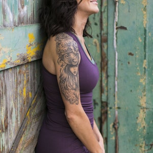 May include: A woman with short, dark hair is wearing a purple tank top and has a large tattoo on her upper arm. The tattoo is a black and white image of a woman with long hair and a floral design.