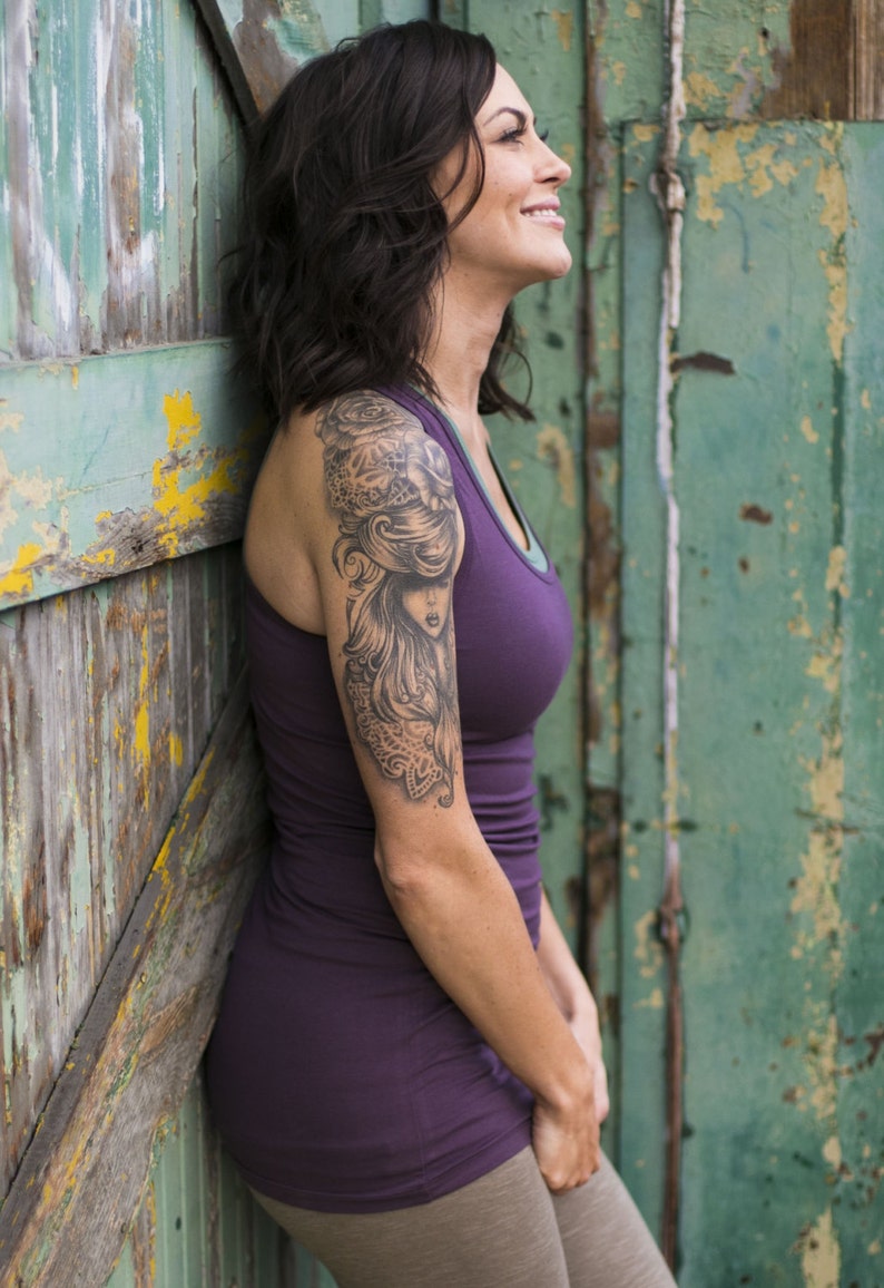 May include: A woman with short, dark hair is wearing a purple tank top and has a large tattoo on her upper arm. The tattoo is a black and white image of a woman with long hair and a floral design.