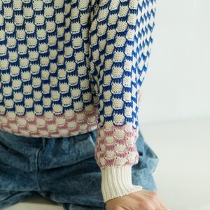 May include: A blue, white, and pink patterned sweater with a unique design. The sweater features a repeating pattern of blue shapes on a white background, with pink accents on the cuffs. The sleeves are long, and the cuffs are ribbed.