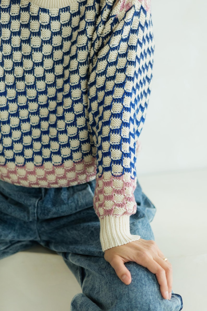 May include: A blue, white, and pink patterned sweater with a unique design. The sweater features a repeating pattern of blue shapes on a white background, with pink accents on the cuffs. The sleeves are long, and the cuffs are ribbed.