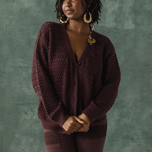 May include: A person wearing a maroon cardigan with a chevron pattern and matching pants. The cardigan has a button closure and the person is wearing gold hoop earrings.