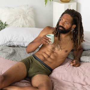 Puede incluir: Una persona con ropa interior verde oliva con una banda negra, tumbada en una cama rosa con una almohada blanca y una planta verde en el fondo.
