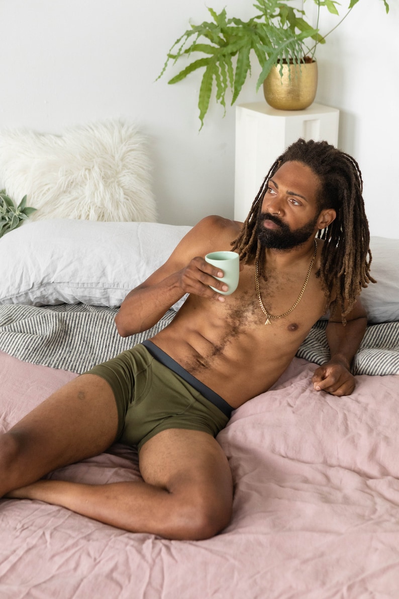 Puede incluir: Una persona con ropa interior verde oliva con una banda negra, tumbada en una cama rosa con una almohada blanca y una planta verde en el fondo.